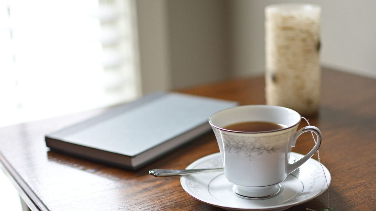 A china teacup and a closed notebook on a dark wooden table by a bright window.