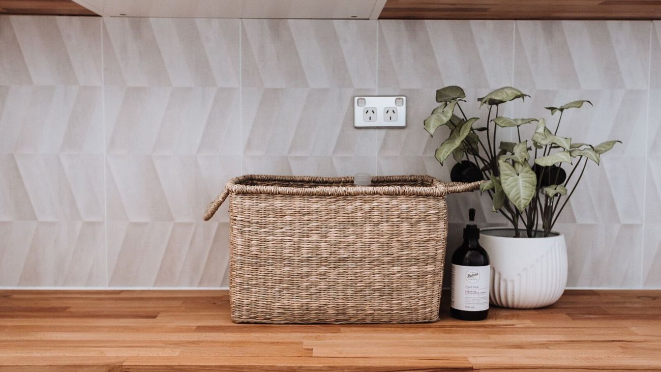 A woven basket and a potted plant on a wooden laundry counter against pale tiles.