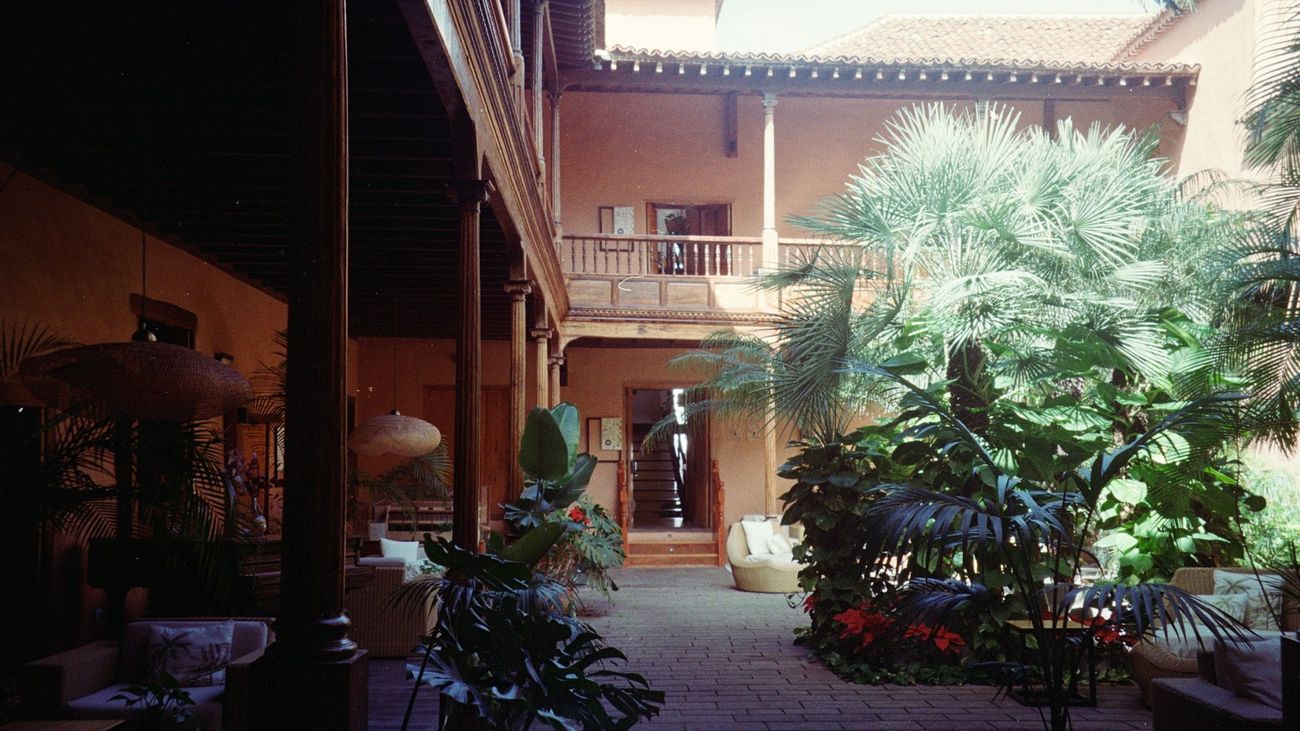 A hotel courtyard with a carved wooden gallery, terracotta walls and palms around a brick-paved path.