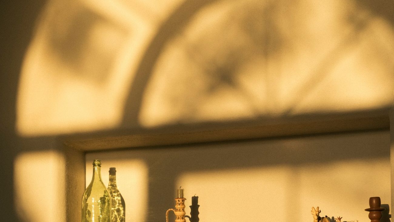 Late sun throwing the shadow of an arched window across a cream plaster wall above a wooden shelf.