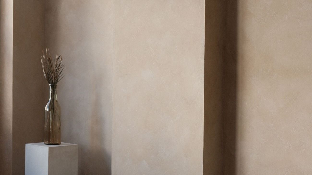 A tall cream plaster arch beside a white plinth holding a vase of dried grass.