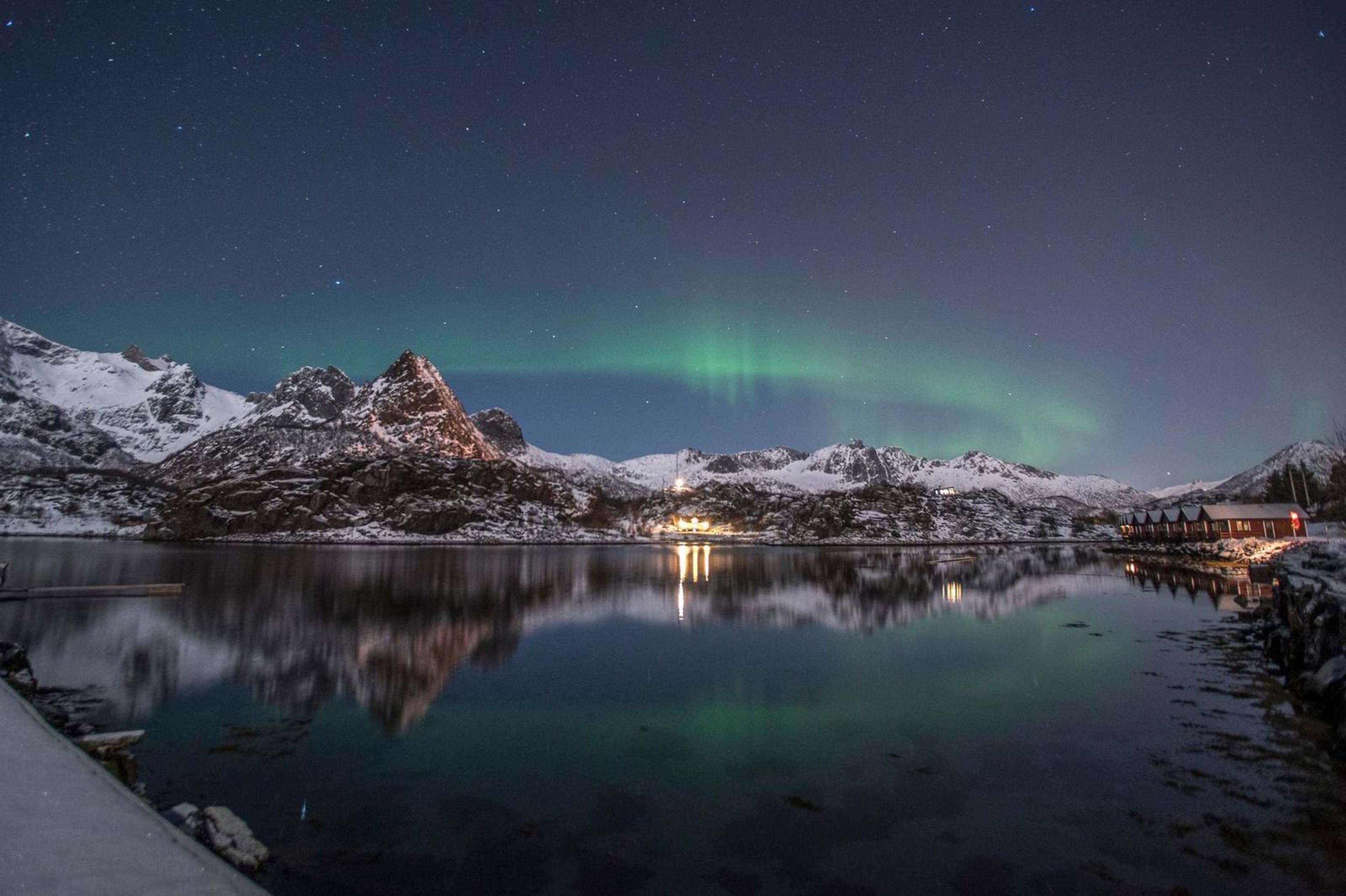 Aurora boreal verde sobre cabañas rorbu rojas reflejadas en un fiordo de las Lofoten