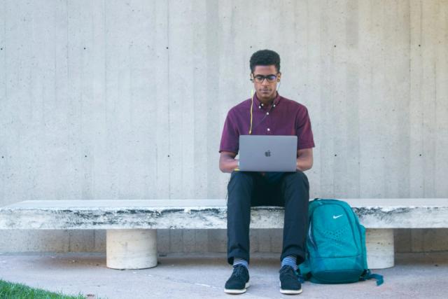 A student sitting on a bench with a laptop on his lap and a backpack beside him.