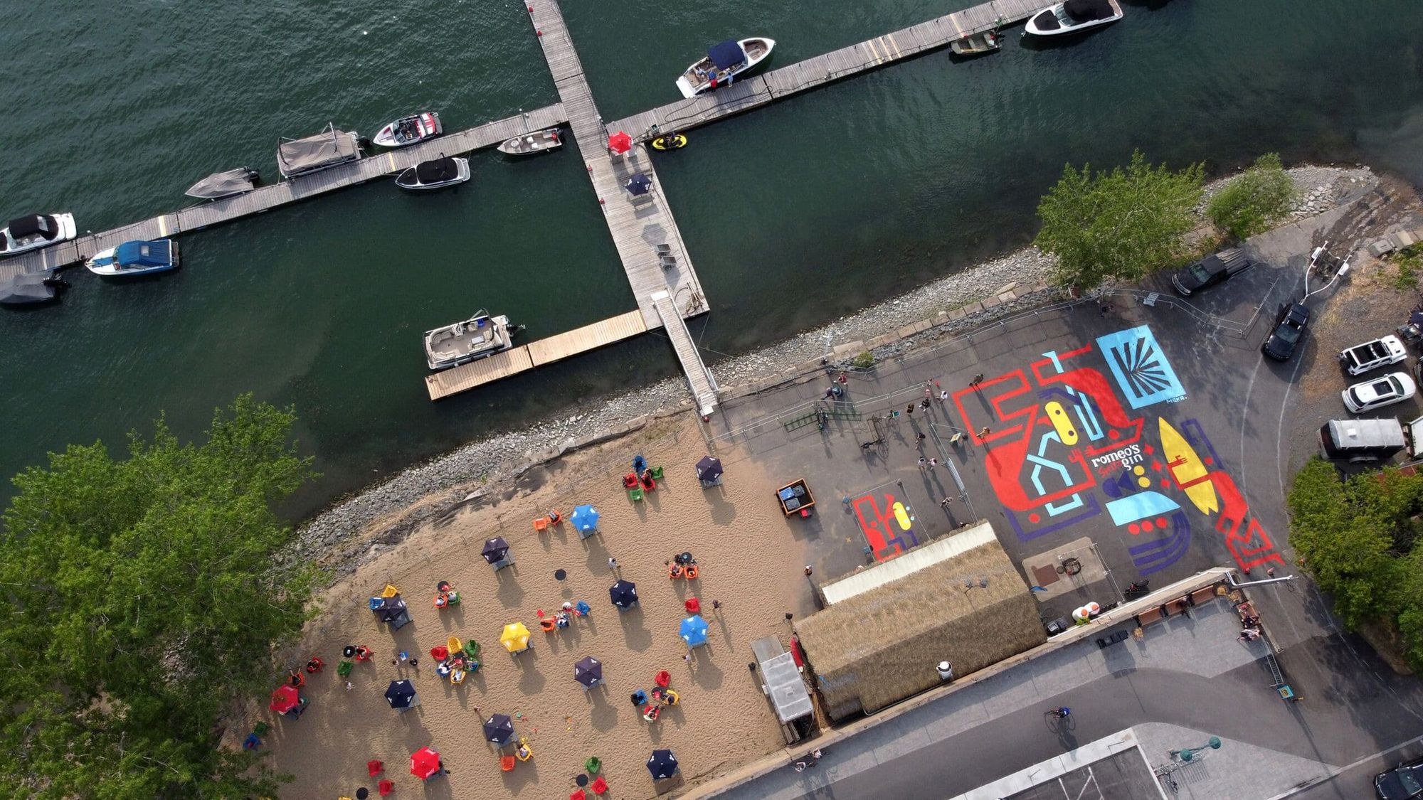 Aerial view of the Romeo's Gin ground mural covering the marina parking lot in Boucherville, crowd on the beach beside it