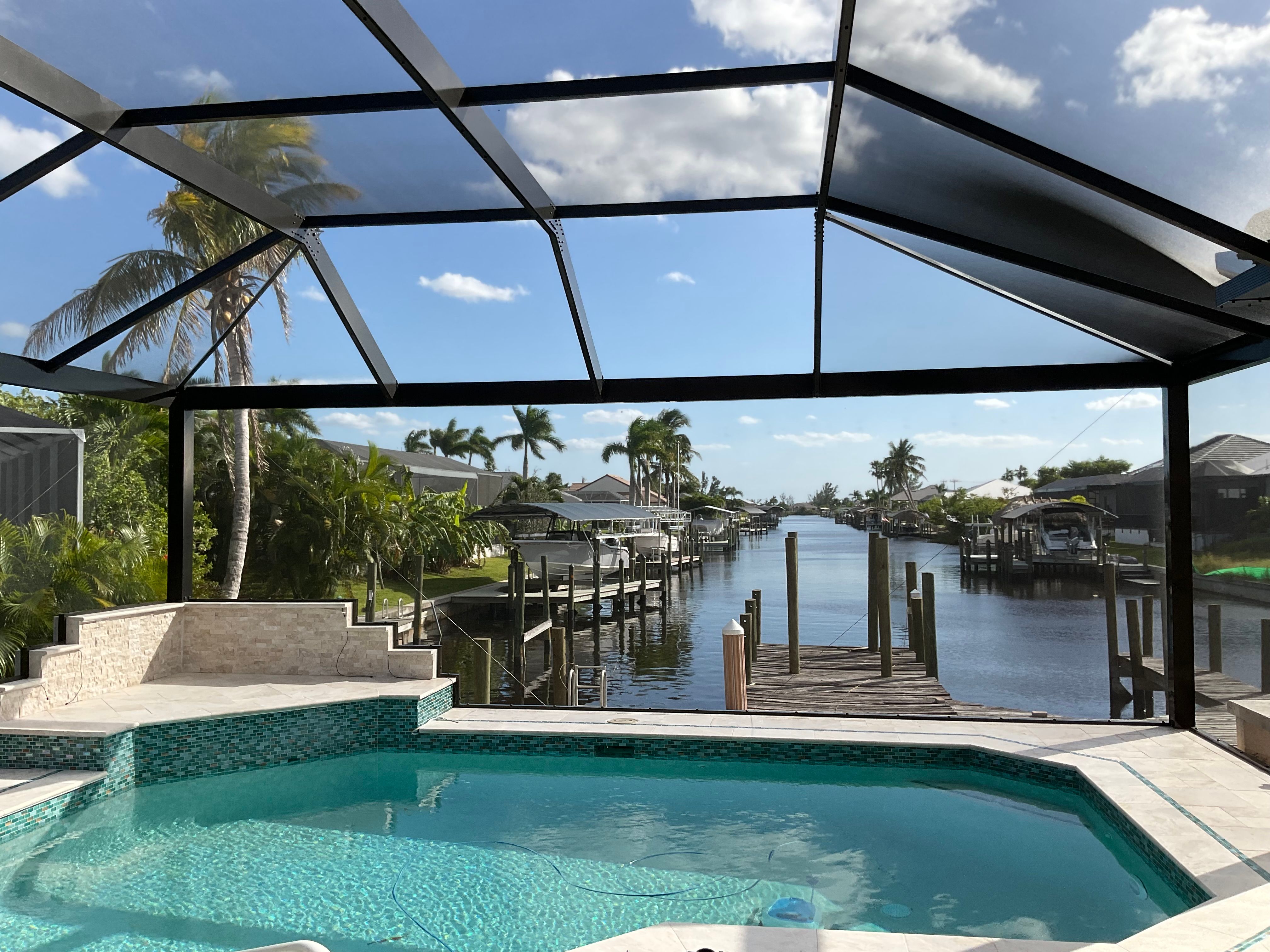 Dark bronze aluminum pool screen enclosure interior, freeform pool with elevated spa, waterfront canal view with boat docks and palm trees, South Florida — AB Aluminum & Screens