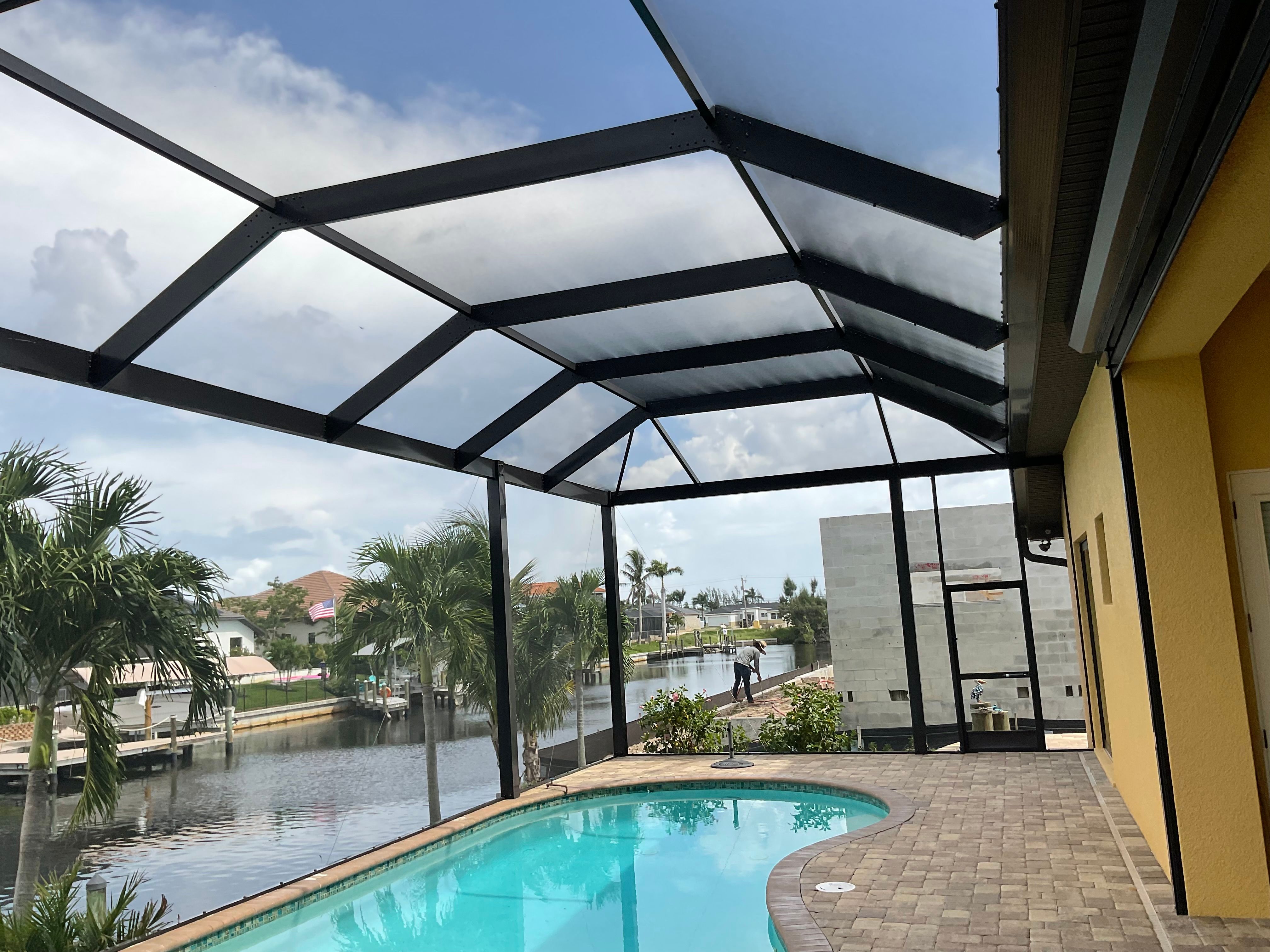 Dark bronze aluminum pool enclosure interior with curved pool and paver deck overlooking wide waterfront canal with docks, palm trees and luxury homes — AB Aluminum & Screens