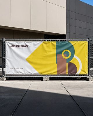 Witte werfbanner met aan de rechterkant een gele driehoek die wijst naar links. op het gele vlak staat het logo symbool van Atelier Buyck. Aan de linkerkant langs de bovenkant van de banner staat de naam voluit geschreven onderaan de linkerkant staat de link naar de website.
