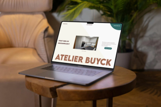 Een Laptop in een living, een zetel en een kamerplant in de achtergrond. De laptop staat op een tafeltje met een metalen onderstel. De laptop toont de website van atelier buyck: https://atelierbuyck.be