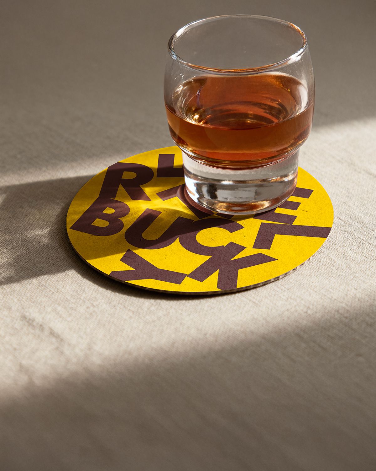 Onderlegger voor drank mockup in het geel met daarop de letters van Atelier Buyck in het donkerbruin willekeurig verspreid over het oppervlak.