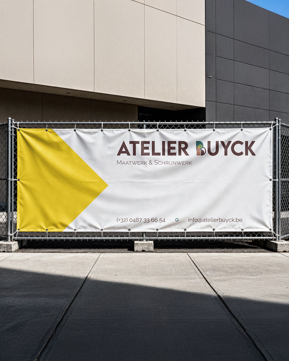 Witte werfbanner met aan de linkerkant een gele driehoek die naar rechts wijst. Aan de rechterkant van de banner staat het volledige logo: de naam Atelier buyck met de b vervangen door het logo symbool, onderaan de rechterkant staat de informatie: telefoon en email