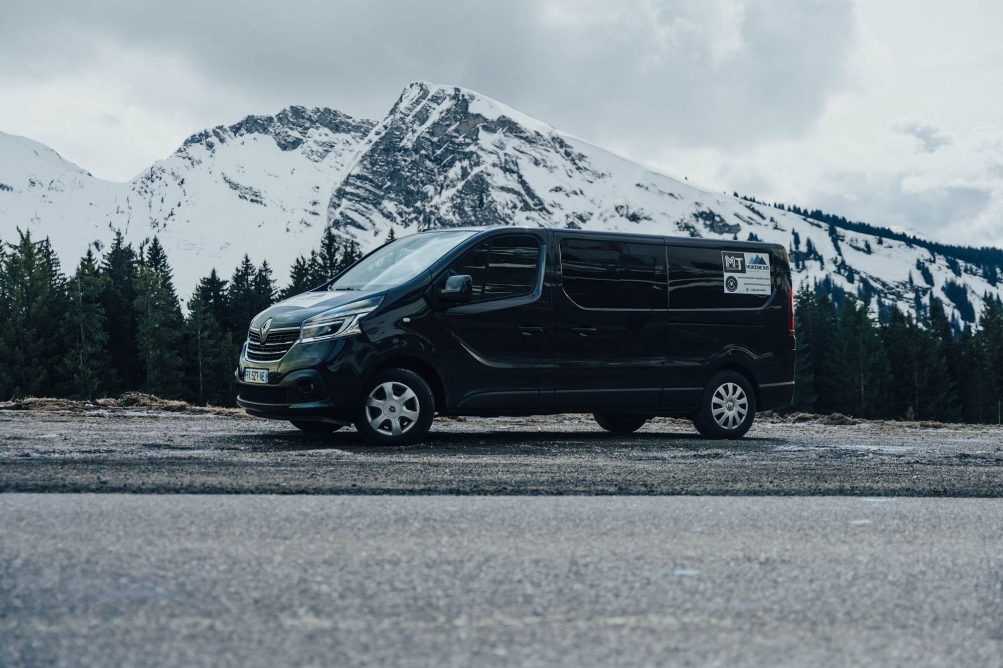 Black Renault Trafic 8-seat minibus side on, with snow-capped mountains behind