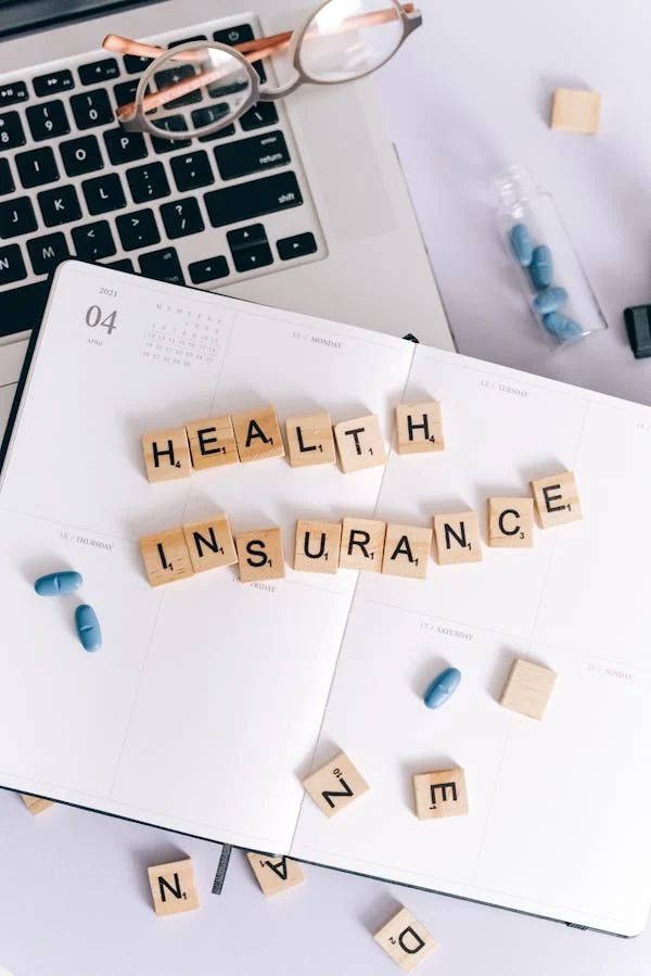 Scrabble tiles spelling "HEALTH INSURANCE" on an open planner, with blue pills, eyeglasses, and a laptop.