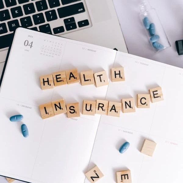 Scrabble tiles spelling "HEALTH INSURANCE" on an open planner, with blue pills, eyeglasses, and a laptop.