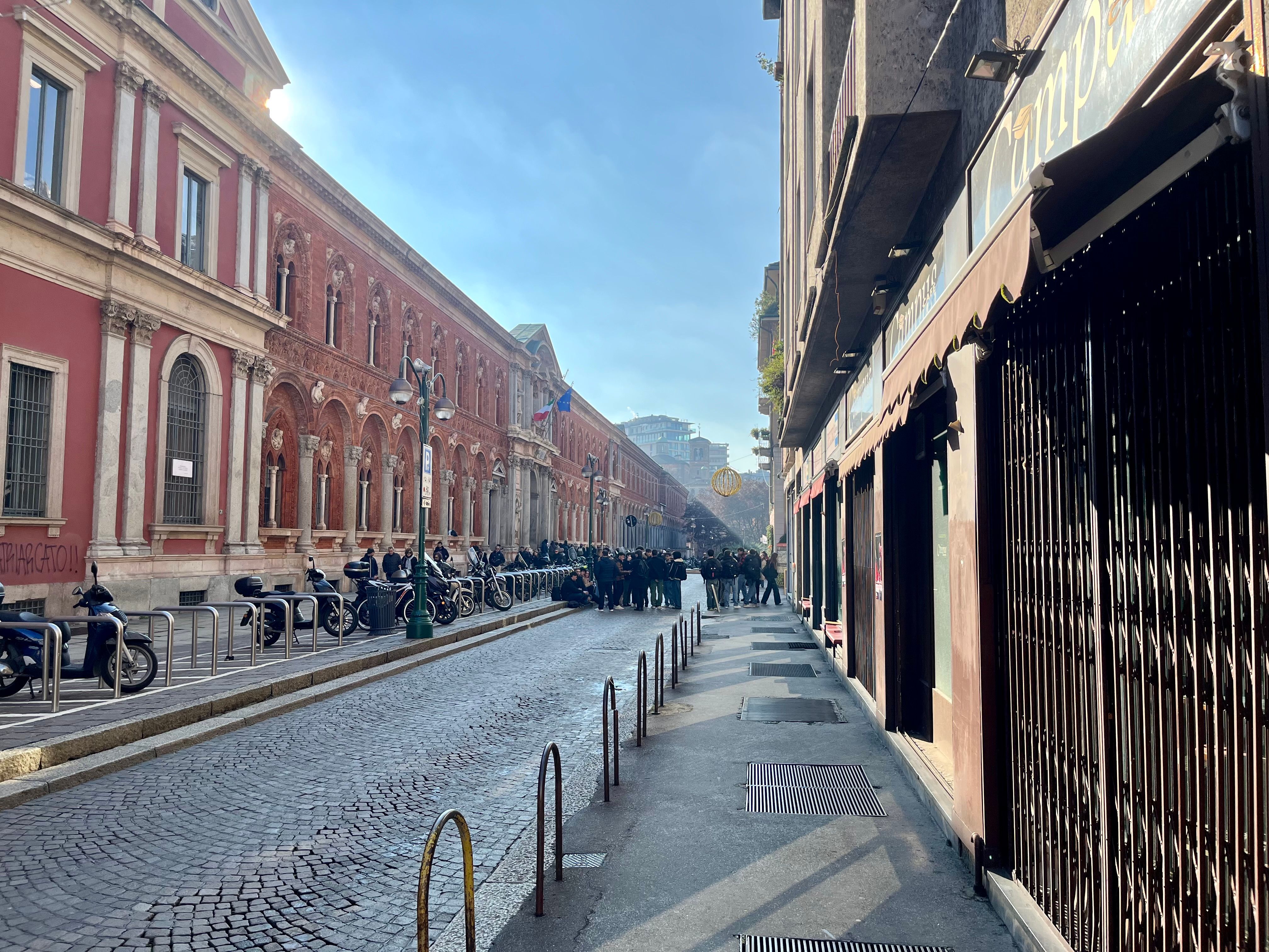 Facade of a bar facing Milan's main road, closed shutters, closed storefront windows, closed premises, sidewalk, cobblestone paving, buildings with windows and balconies