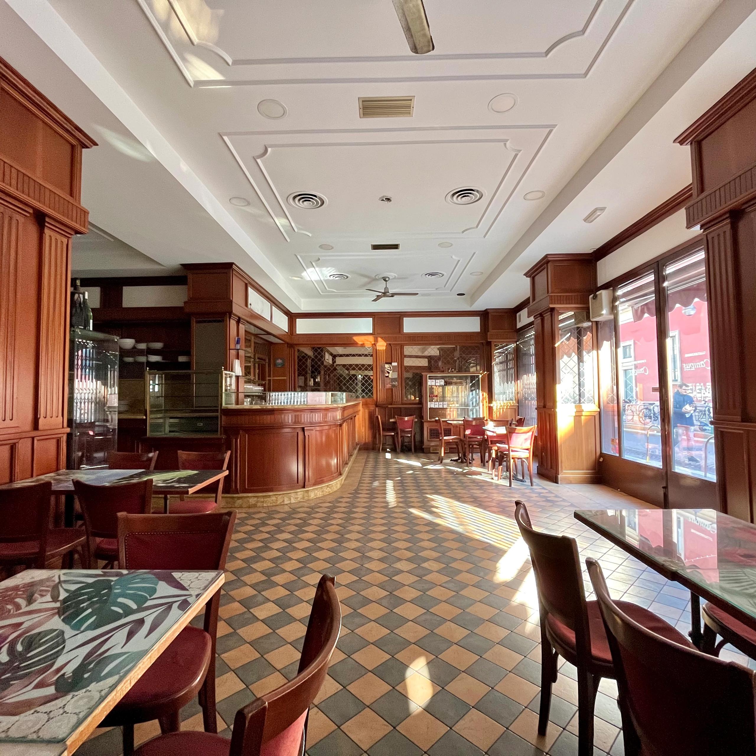 interno del bar, con bancone, vetrina, tavolini e sedie, specchi a parete, pale per ventilazione sul soffitto