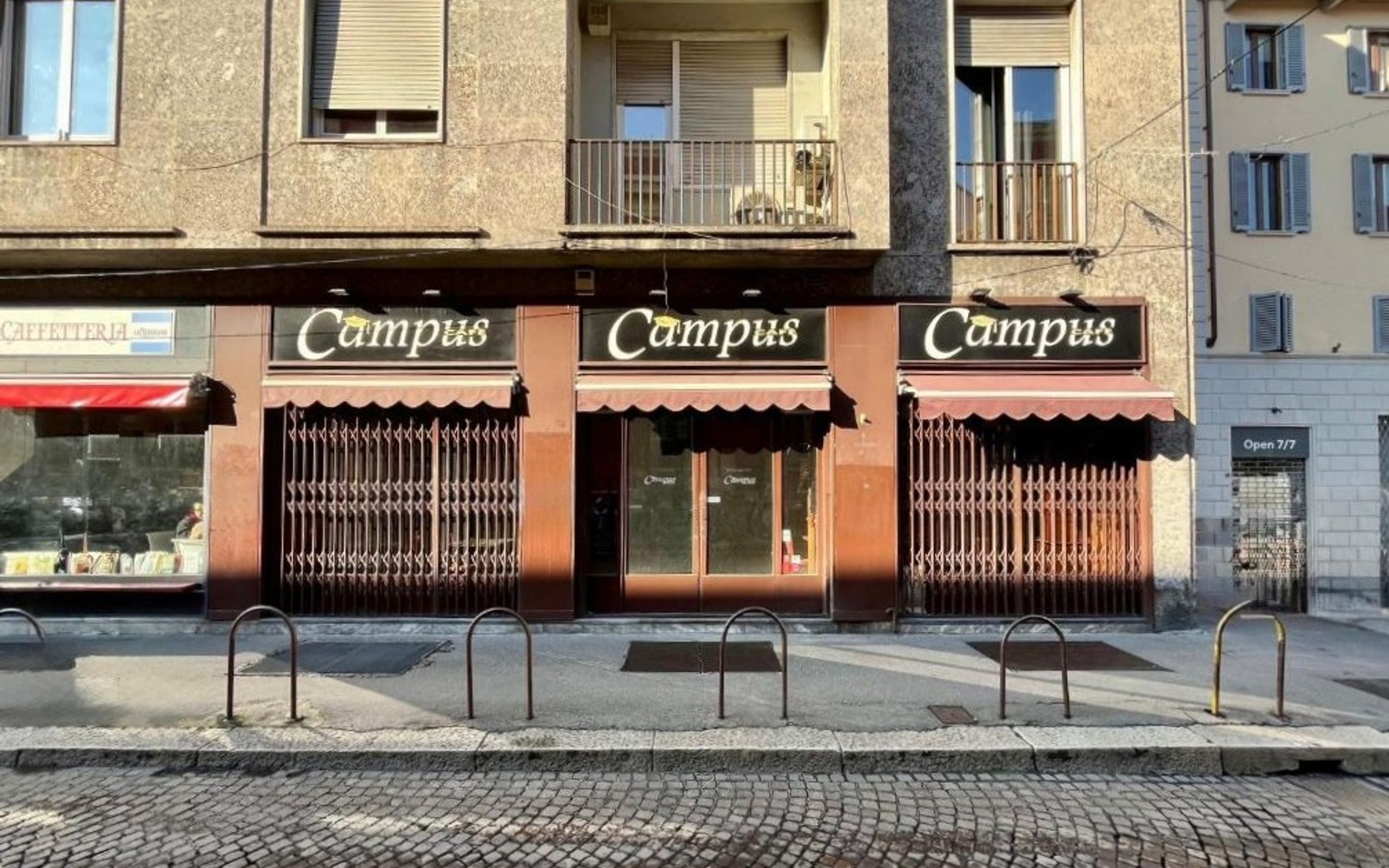 facciata di un bar di fronte alla statale di Milano, saracinesche chiuVetrine chiuse, locale chiuso, marciapiede, pavè, edifici con finestre e balconi
