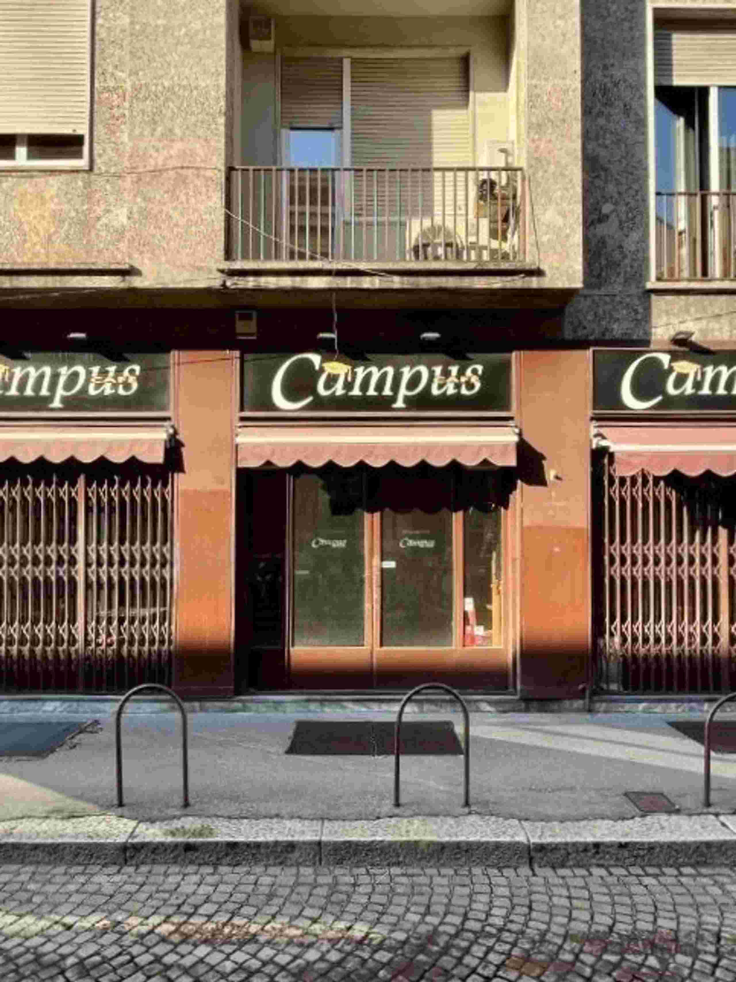 facciata di un bar di fronte alla statale di Milano, saracinesche chiuVetrine chiuse, locale chiuso, marciapiede, pavè, edifici con finestre e balconi