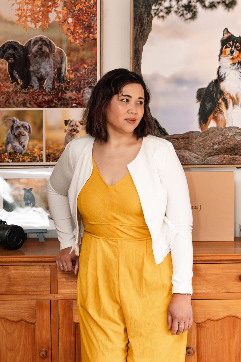Pet photographer Ina J in a yellow jumpsuit standing beside her camera in her studio, large framed dog portraits behind her