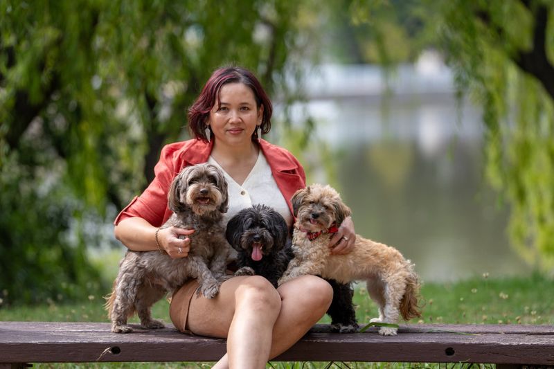 Pet photographer Ina J in a rust jacket sitting on a park bench by the water with three small dogs on her lap