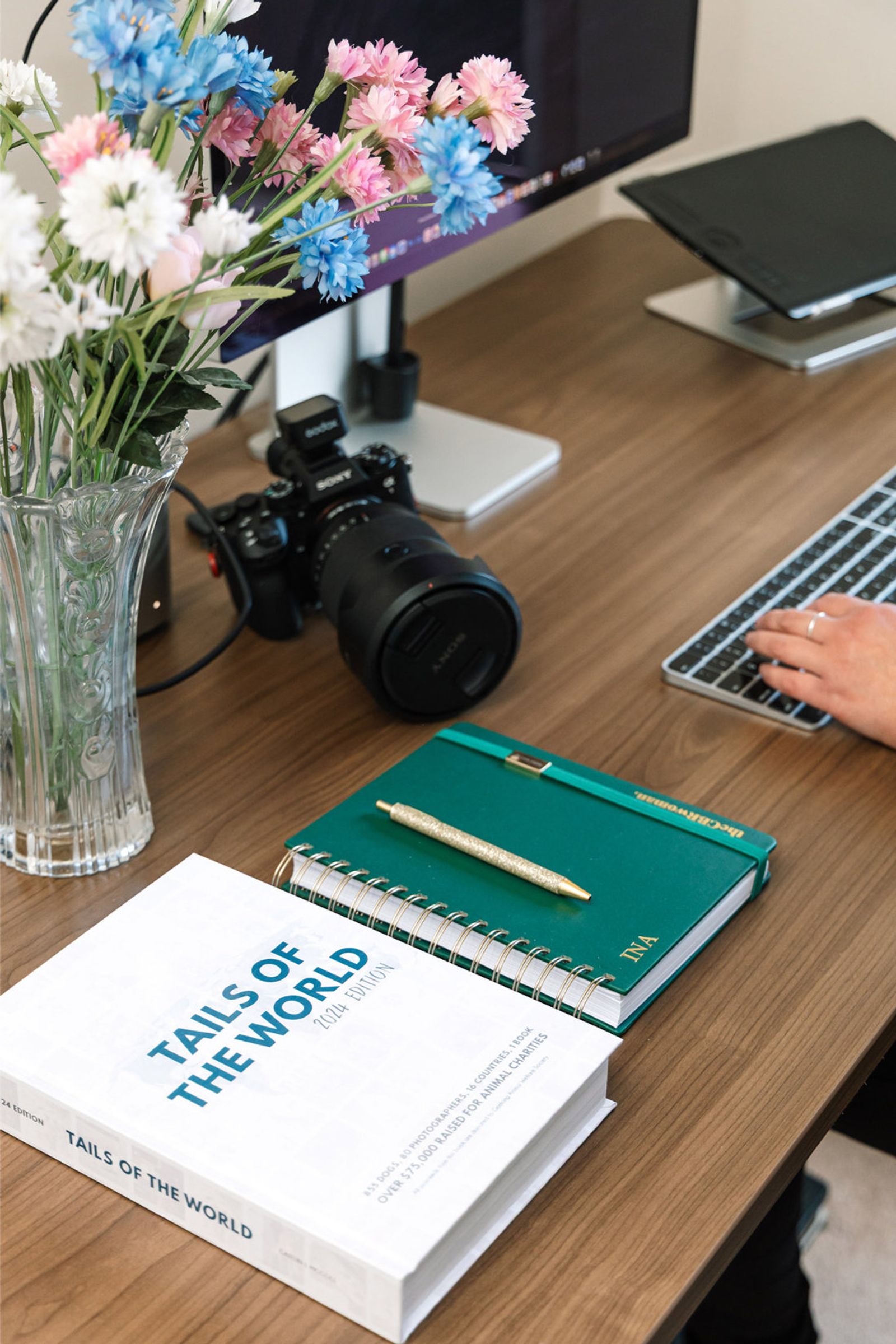 A desk flatlay with a Sony camera, spring flowers, a personalised notebook and a Tails of the World charity book