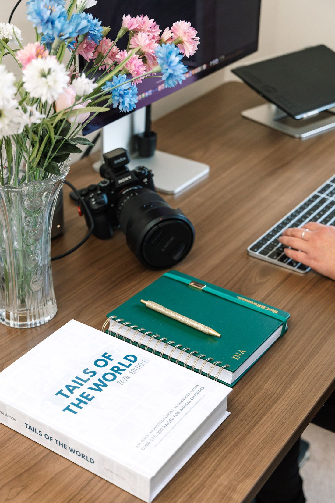 A desk flatlay with a Sony camera, spring flowers, a personalised notebook and a Tails of the World charity book