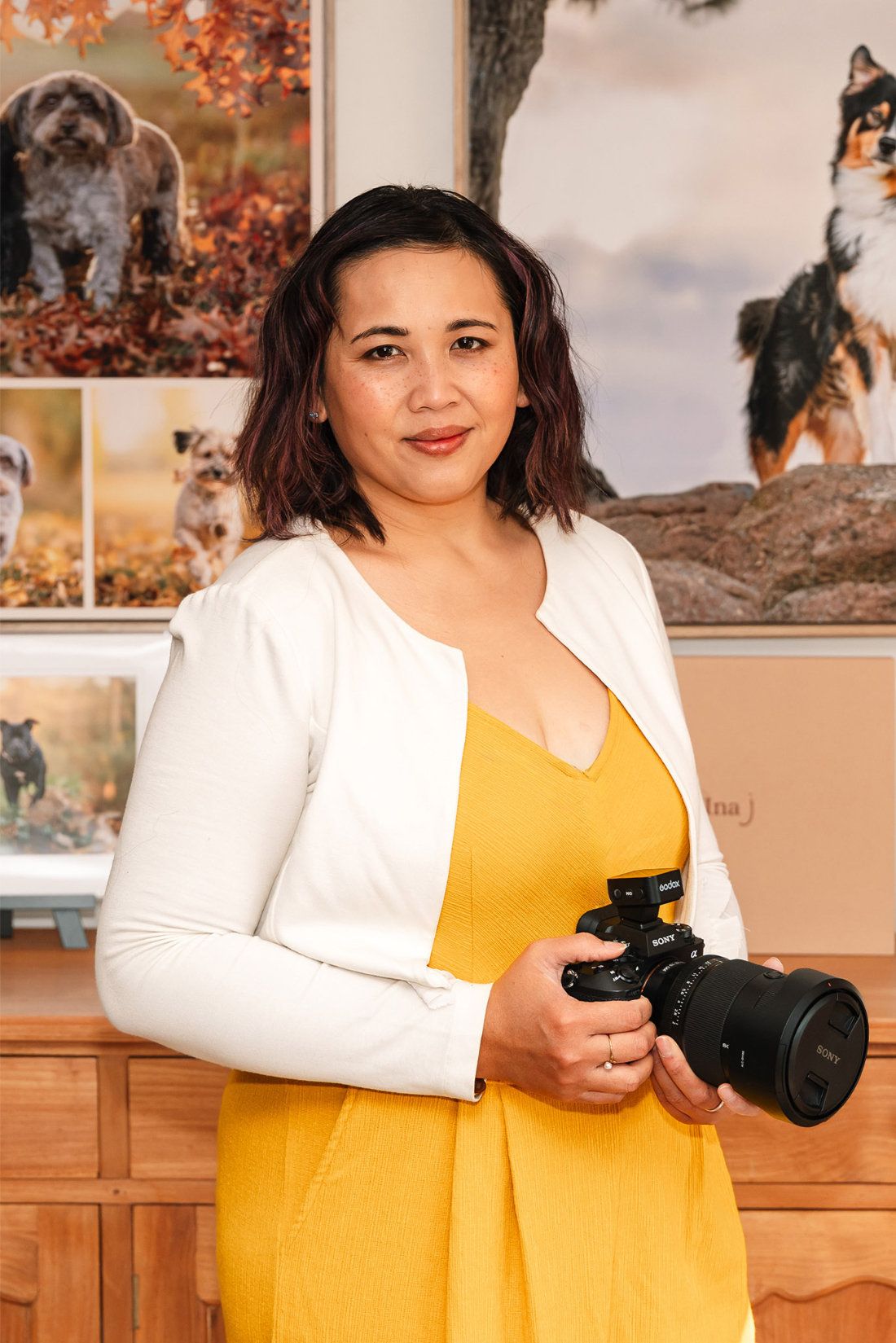 Pet photographer Ina J in a yellow dress holding her Sony camera in the studio, framed dog portraits behind her