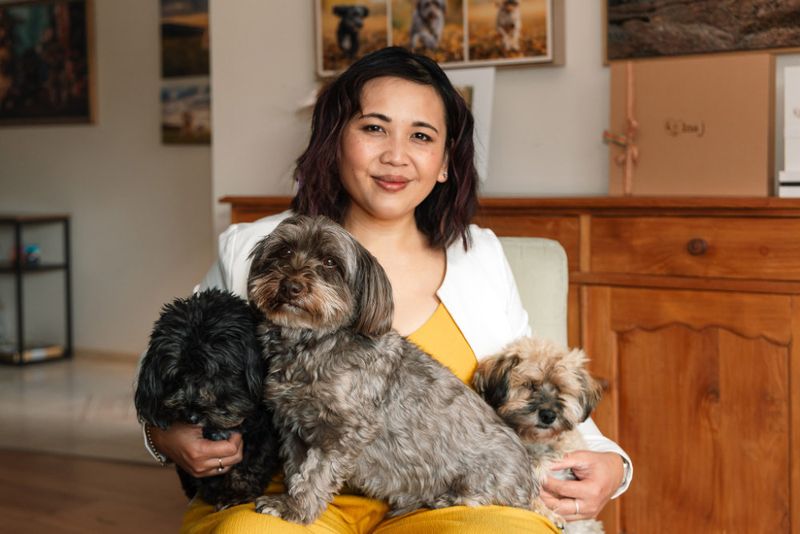 Pet photographer Ina J in a white cardigan seated with three small dogs on her lap in her studio