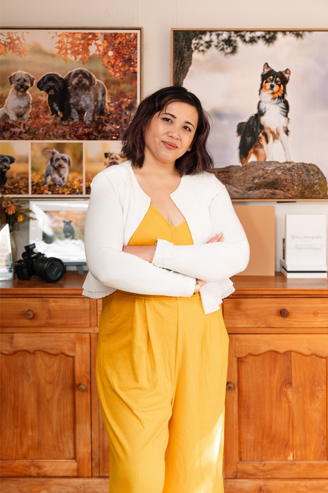 Pet photographer Ina J standing with her arms crossed in front of large framed dog portraits in her studio