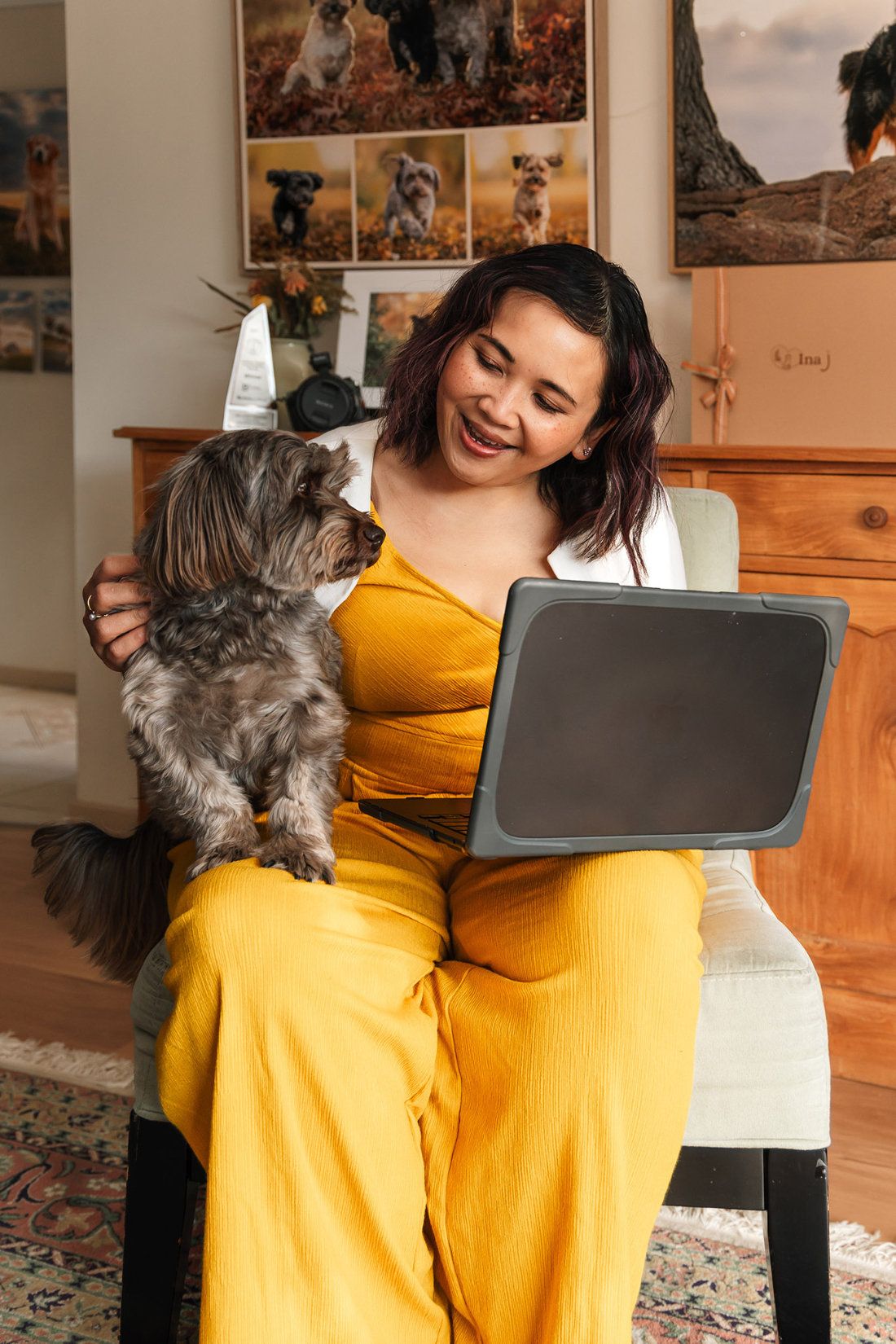 Pet photographer Ina J in a yellow jumpsuit on a chair with a small grey dog nuzzling her, a tablet on her lap