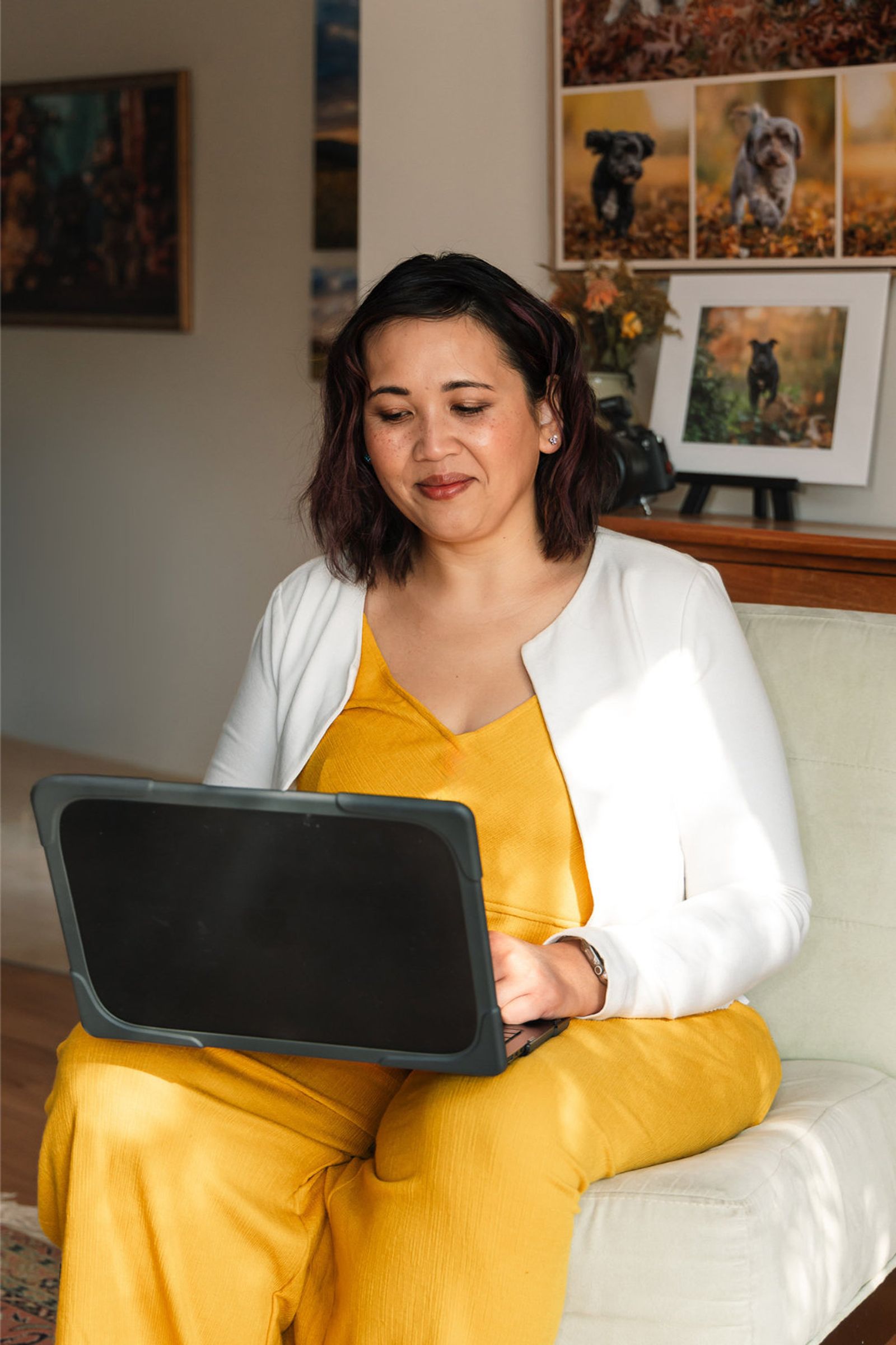 Pet photographer Ina J in a yellow jumpsuit working on a tablet on her studio sofa, framed dog portraits behind her