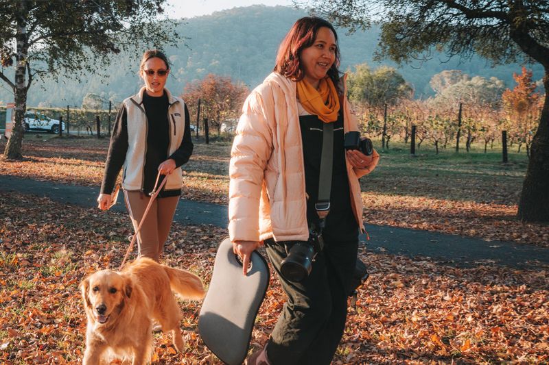 Pet photographer Ina J in a cream puffer jacket carrying camera gear, walking a golden retriever with a client along a leaf-covered vineyard path in autumn