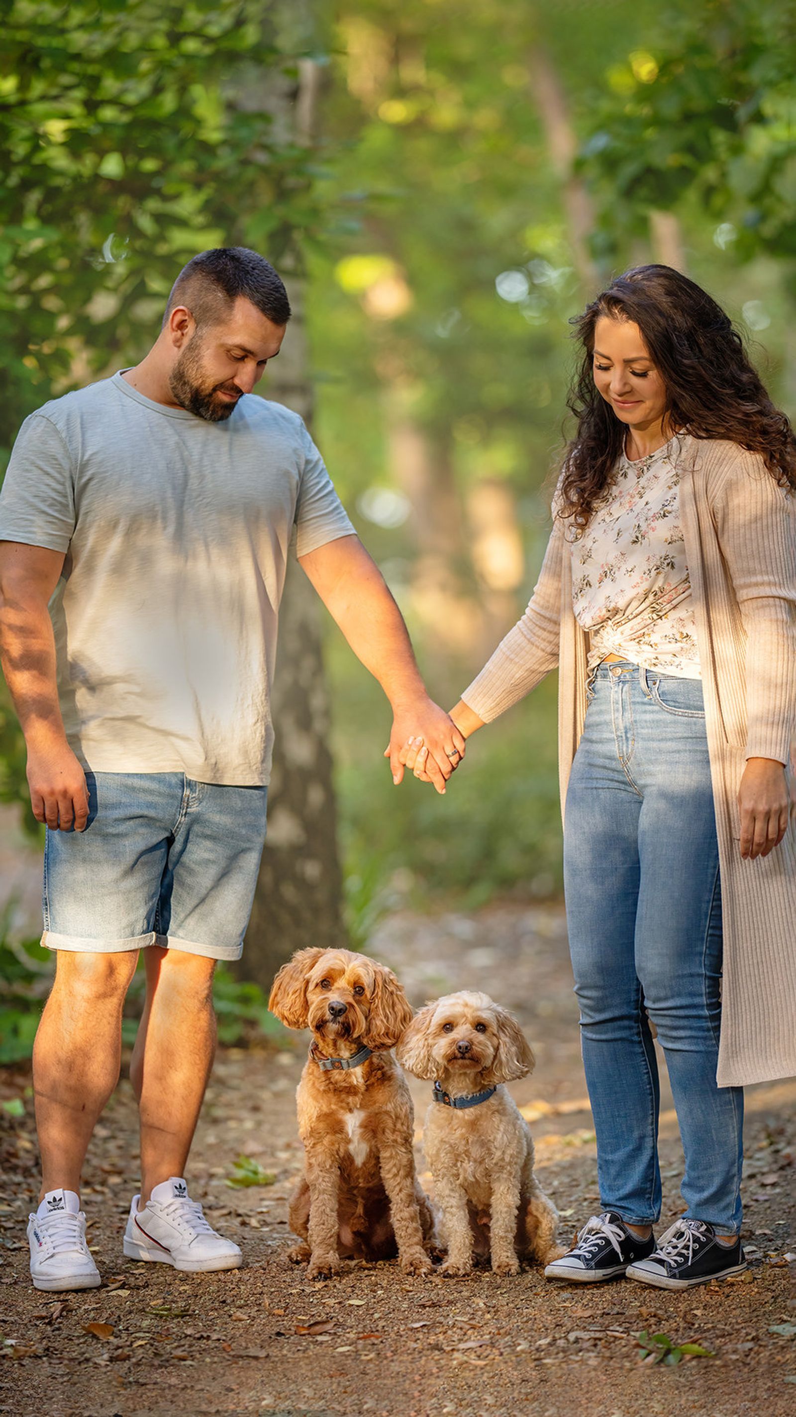 A couple holding hands on a woodland path with their two apricot cavoodles sitting at their feet