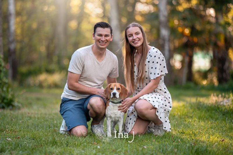 A smiling young couple crouched together in a sunlit park with their beagle sitting between them