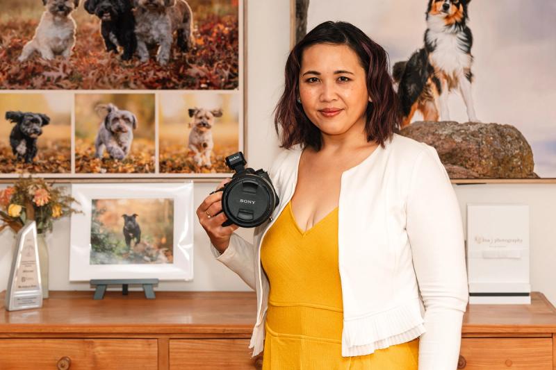 Pet photographer Ina J in a yellow jumpsuit holding a Sony camera, standing by pet photos on a wall in her Canberra studio Ina J Photography a pet photography business coach and mentor