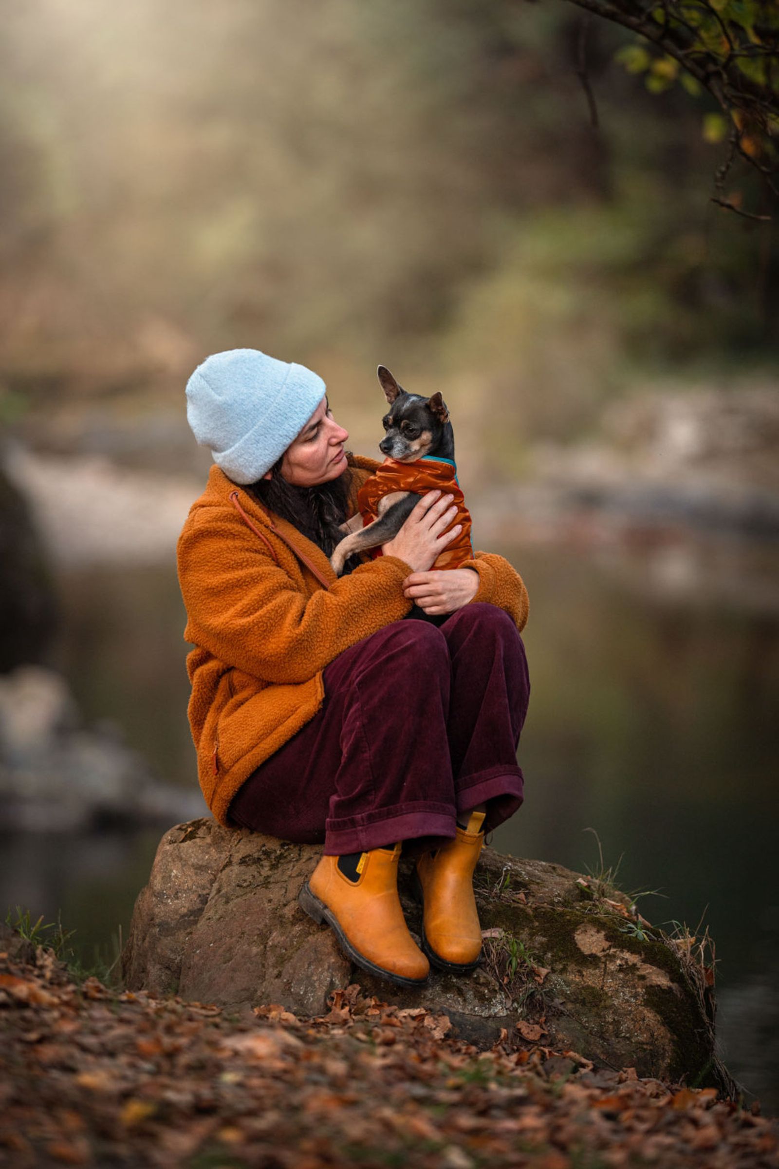 A woman in an orange jacket and blue beanie cuddling a small chihuahua on a rock by a river in autumn
