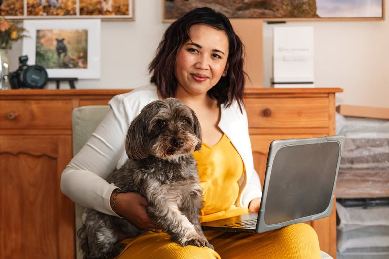 Pet photographer Ina J in a white blazer sitting in her studio with a laptop and a small grey terrier-cross dog on her lap, framed dog portraits behind her