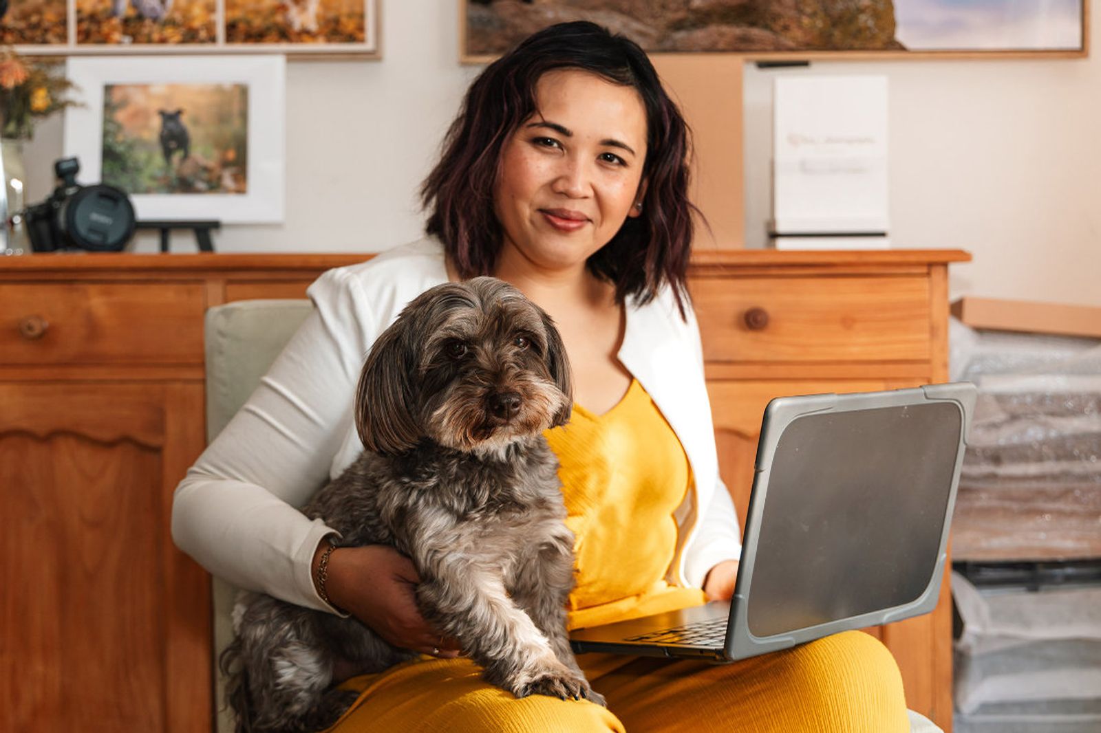Pet photographer Ina J in a yellow jumpsuit sitting with a small grey dog on her lap and a laptop, framed prints behind her