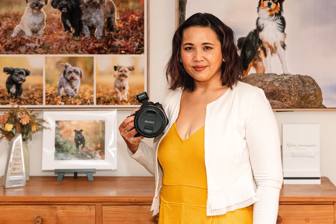 Pet photographer Ina J in a yellow jumpsuit holding her Sony camera in the studio in front of framed prints