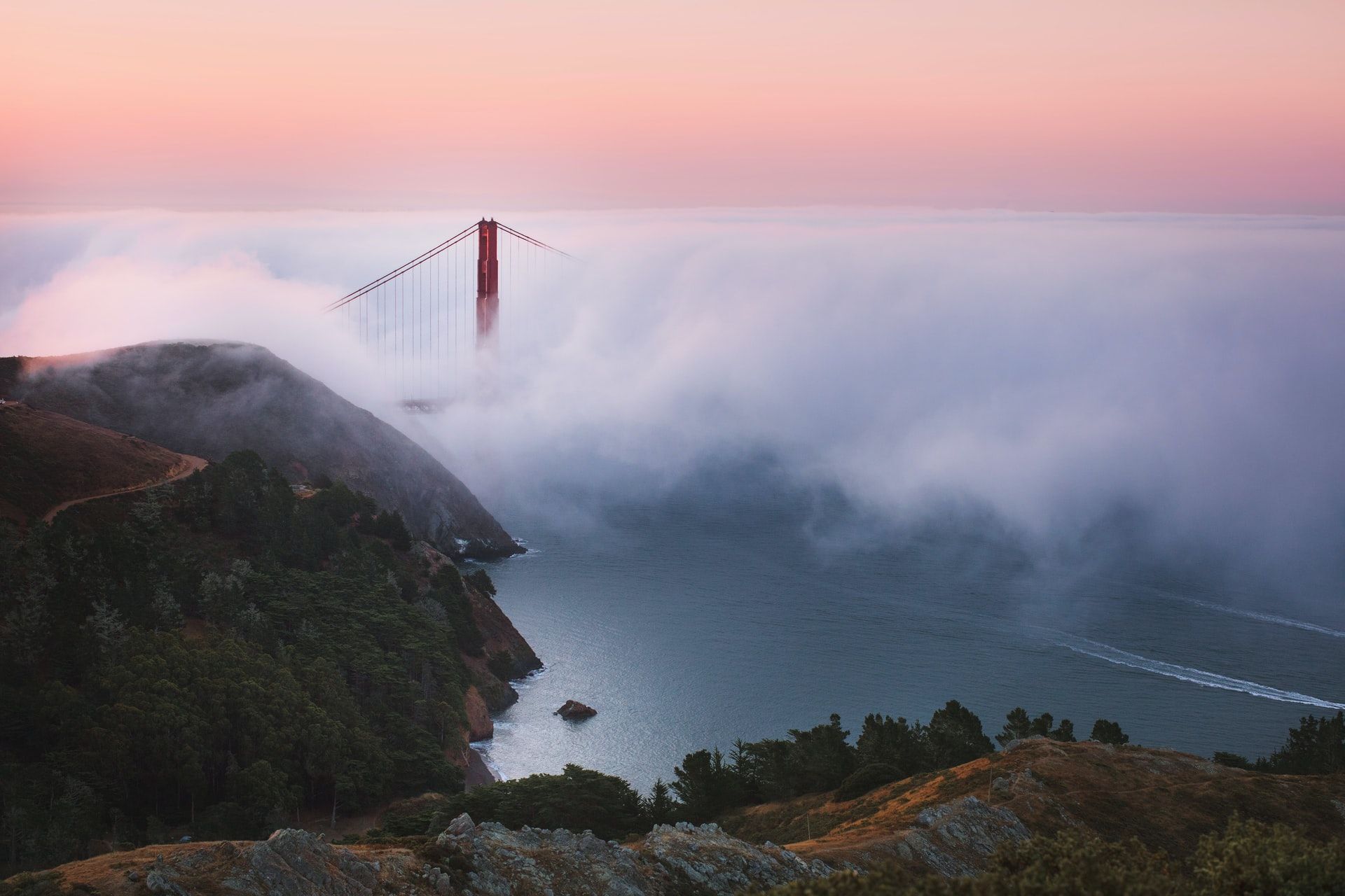 The Golden Gate Bridge partially shrouded in fog at sunset, with the bay and surrounding hills visible.