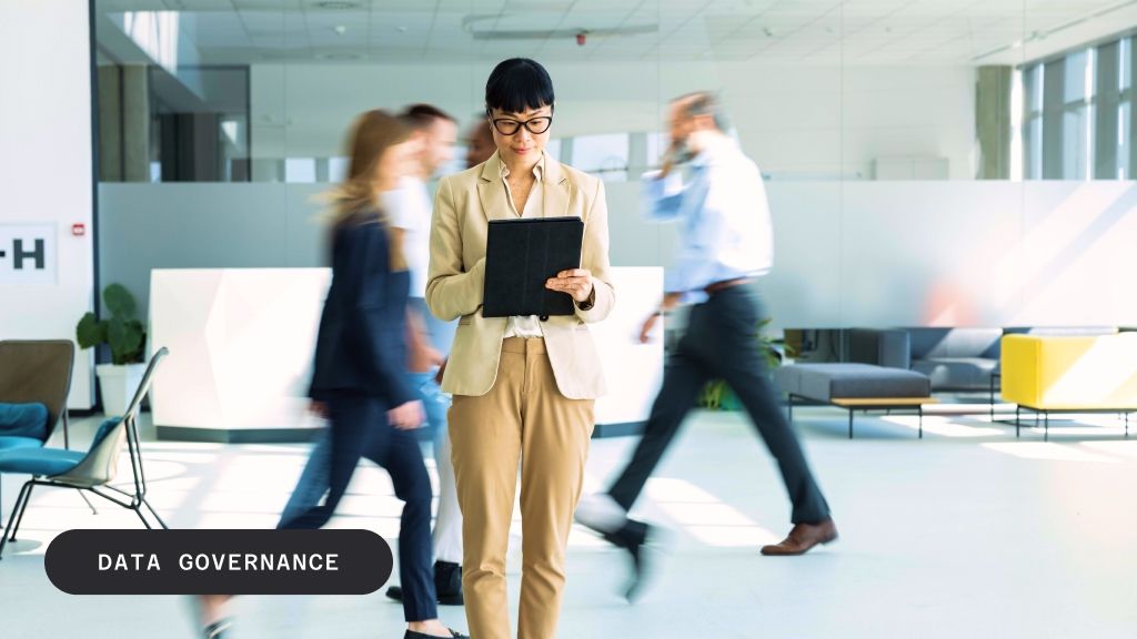 A woman in a blazer and trousers looks at a tablet in a modern, busy office environment with blurred people walking in the background. The text "DATA GOVERNANCE" is overlaid in the bottom left.