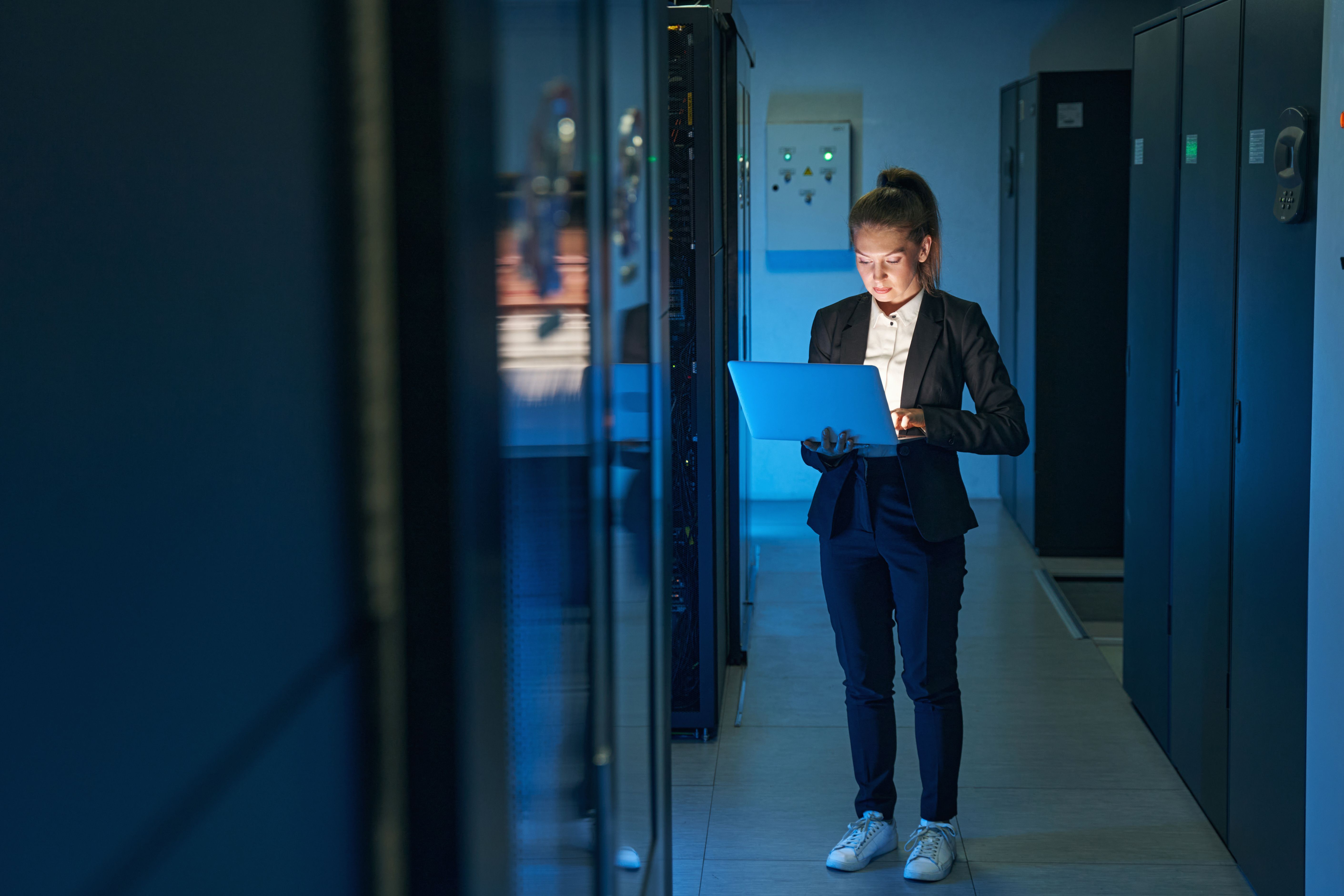 A woman in a dark server room stands focused on a laptop, illuminated by its light, wearing a dark suit and sneakers.