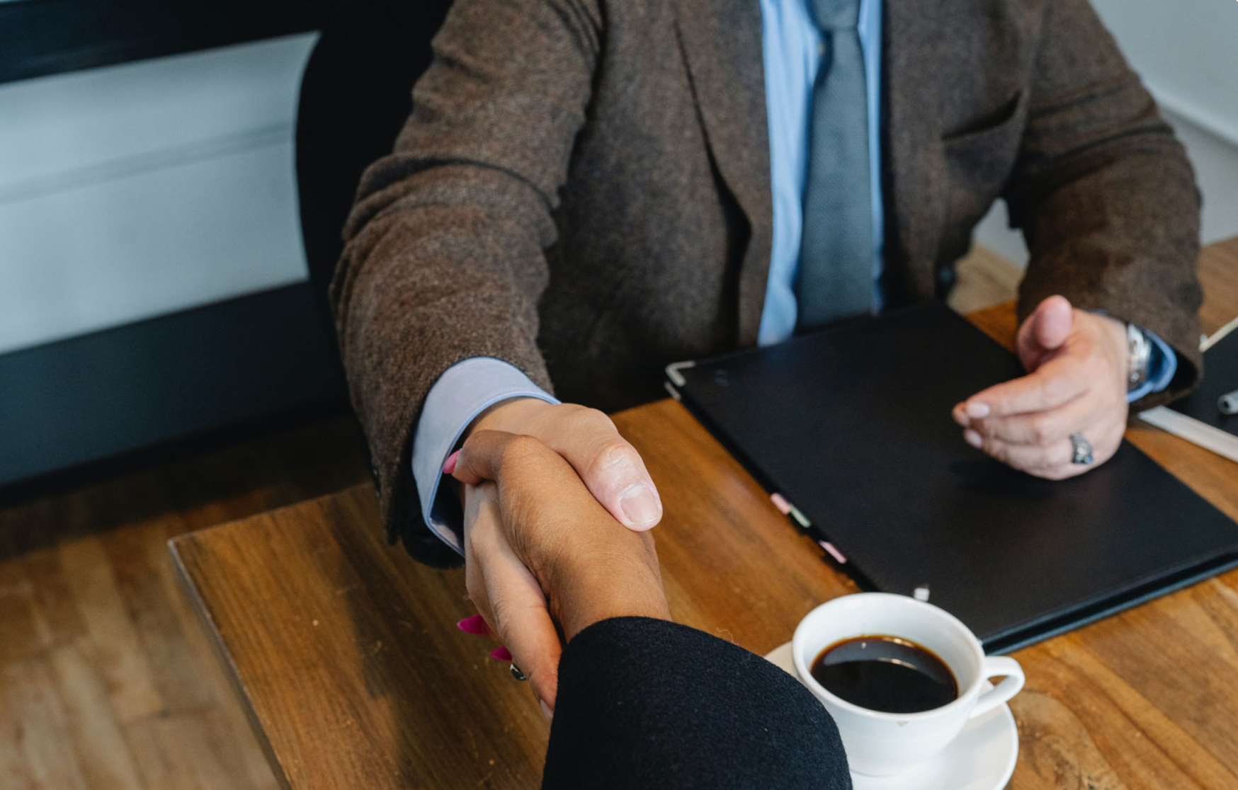 A photograph of two people shaking hands over a table, with a coffee in shot.