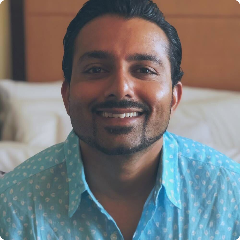 A person with short dark hair and a beard is smiling while wearing a light blue patterned shirt, with a neutral-colored headboard and bed in the background.