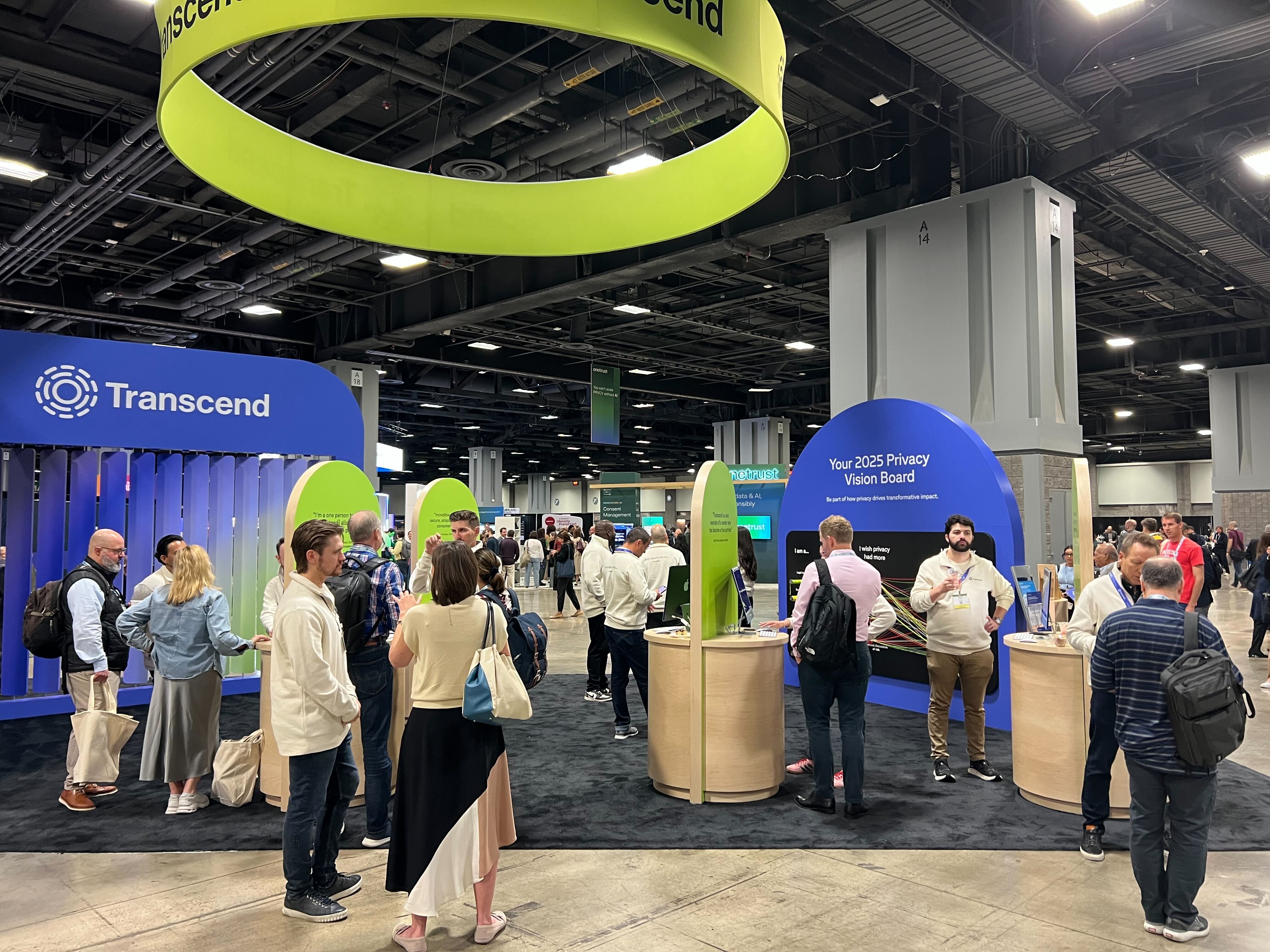 A busy exhibition hall with attendees interacting at various booths, including a large blue privacy vision board and a green Transcend booth hanging above. People are engaged in conversations and exploring different displays.