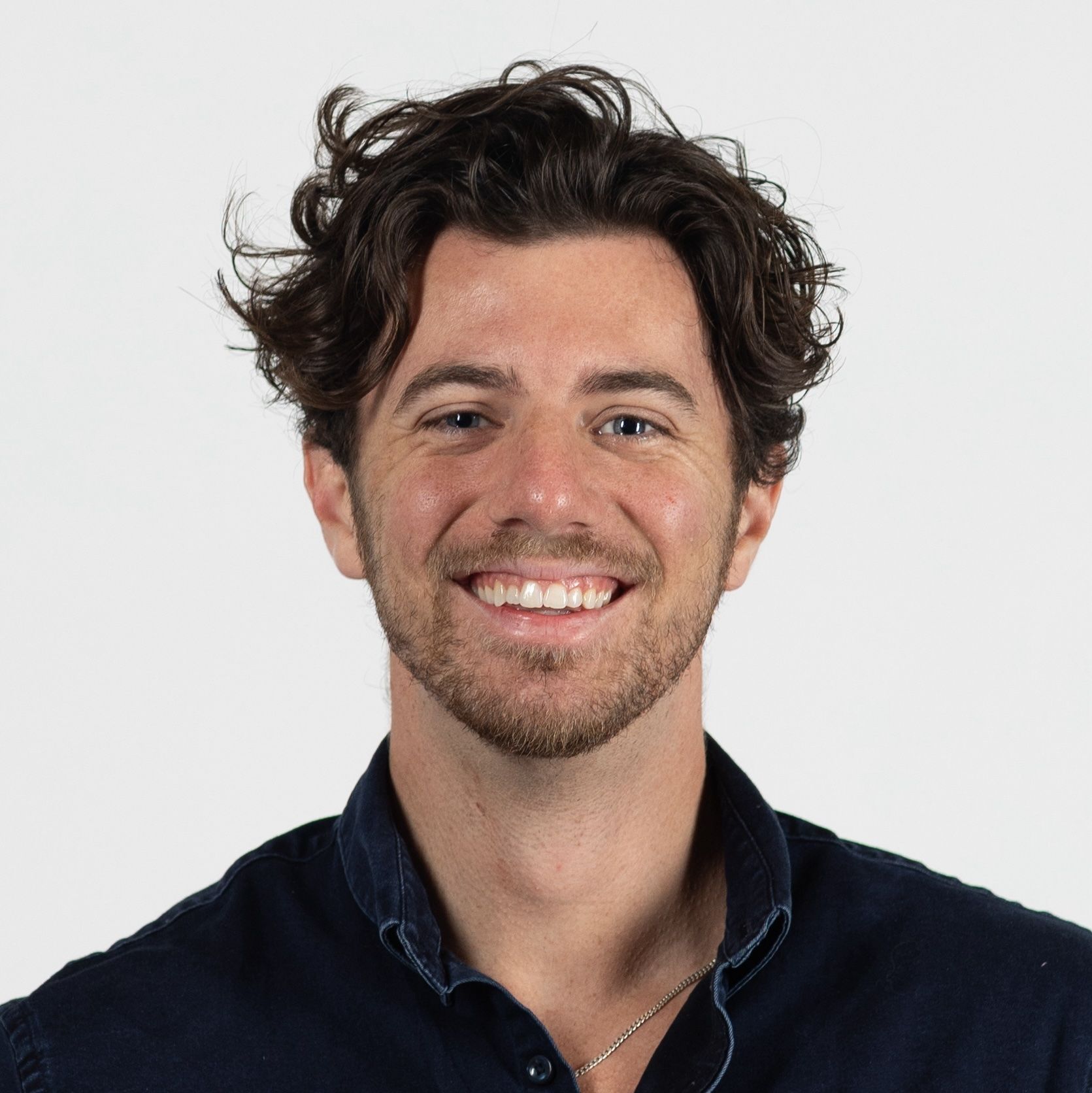 A man with curly dark hair and a beard smiles, wearing a dark collared shirt against a plain white background.