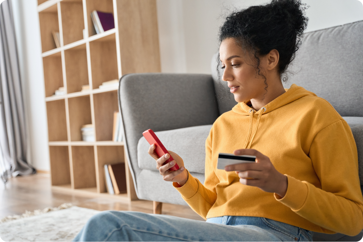 Young woman on floor of apartment leaned up against couch holding credit card and smartphone