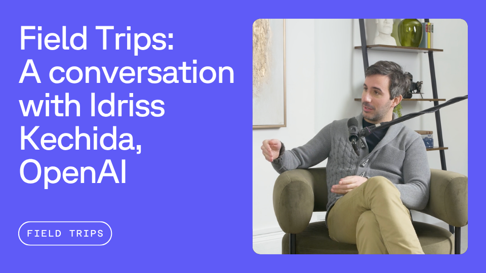 A man seated in a chair speaking into a microphone, wearing a gray sweater and khaki pants. A text overlay reads, "Field Trips: A conversation with Idriss Kechida, OpenAI."