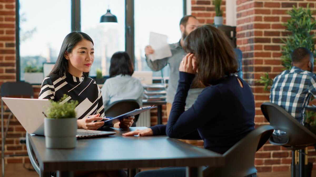 A photograph of two women having a discussion at a table in a modern office setting, with other colleagues working in the background.
