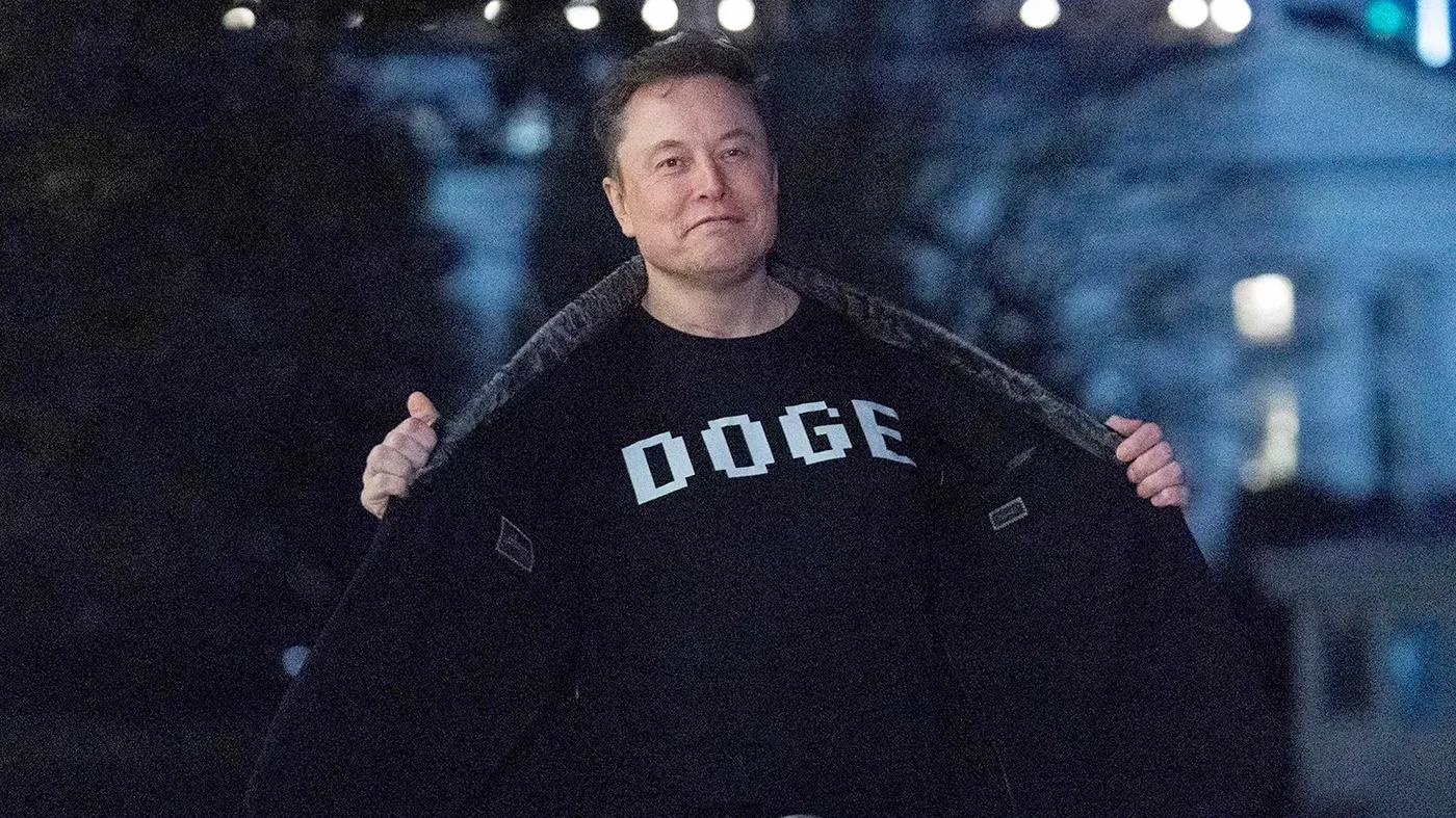 A person stands outdoors at night, holding open a jacket to reveal a black shirt with the word "DOGE" printed on it in bold white letters.