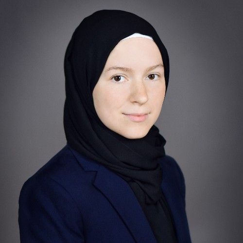 A person wearing a black headscarf and a navy blazer, facing the camera against a gray background.